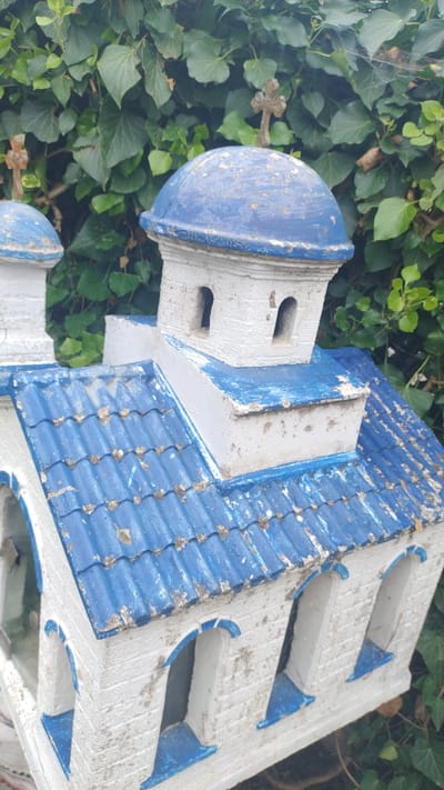 This is a close-up, outdoor, eye-level shot of a white miniature church model with blue-painted details, set against a backdrop of lush green ivy foliage in Νέα Κερασιά, Greece. The model features a blue tiled roof with white trim, and several white walls with arched blue window openings. Above the main structure, two white tiered towers are topped with blue domes. The larger dome on the right has a weathered, antique-looking brown cross affixed to its peak. The smaller dome on the left, partially visible, also has a similar brown cross on top. The paint on the model is somewhat chipped and aged, suggesting it has been exposed to the elements. The ivy leaves in the background are various shades of green, some slightly browned, indicating natural outdoor conditions. The lighting suggests it is daytime, likely overcast or in shade, given the soft light and absence of strong shadows.