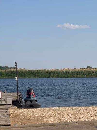 The image depicts a serene waterfront scene on a clear, sunny day in Vodnik, Russia. A calm, deep blue body of water occupies the majority of the frame, with a gentle ripple pattern on its surface. In the foreground, a narrow strip of sandy beach is visible, sparsely covered with light-colored pebbles. To the left, a wooden pier extends into the water, with a jet ski docked at its end. The jet ski is predominantly black and gray with red accents. A metal pole with a light fixture and a white electrical box stands near the pier. Beyond the water, a dense line of tall, dry reeds and green vegetation forms a natural barrier, suggesting a marshy or riverbank environment. The sky is a pale blue, with a single, wispy white cloud drifting lazily across it. The overall atmosphere is one of tranquility and leisure.