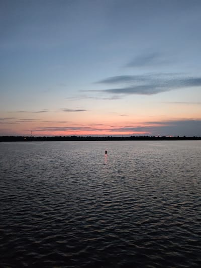 A wide shot captures the Volga River at dusk, near the city of Uglich, Russia. The sky is a gradient of soft blues and pinks, with wispy clouds scattered across the horizon. The water, a deep, dark blue, reflects the twilight sky and is gently rippled. A single red and white buoy stands in the center of the frame, a solitary marker on the vast expanse of water. In the distance, a dark treeline silhouettes the far bank of the river, with faint lights visible indicating the presence of civilization. The overall mood is serene and peaceful, a quiet moment at the close of day.