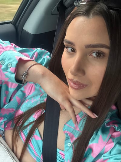 In Draganovtsi, Bulgaria, a young woman with long, dark hair is pictured inside a car. She is wearing a teal and pink patterned shirt and has a silver bracelet on her wrist. Her left hand rests on her chin, and she is looking towards the camera. The interior of the car is visible, including a seatbelt and the car's door panel. The background outside the car appears to be a blurred outdoor scene.