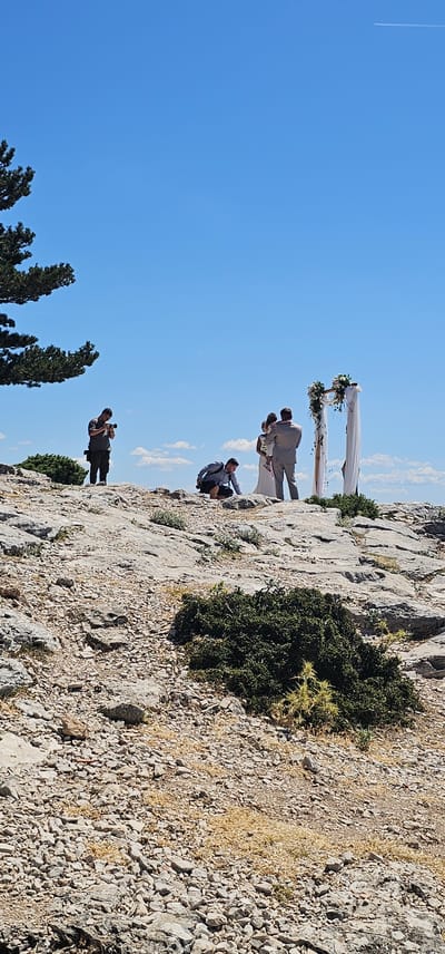 This image captures a wedding ceremony taking place outdoors on a rocky terrain under a clear blue sky with scattered white clouds. The scene is set on a hilltop, likely in the vicinity of Općina Bol, Croatia, given the characteristic rocky landscape and vegetation.
In the center of the frame, a bride in a white wedding dress and a groom in a light-colored suit are standing near a floral archway decorated with white drapery and greenery. The groom is holding a young child.
Two photographers are present, actively documenting the event. One photographer, dressed in a dark shirt and pants, stands to the left of the couple, holding a camera and looking down at it. Another photographer, in a grey shirt and dark shorts, is crouched down in the foreground, closer to the couple, also with a camera.
The environment is dry and arid, with numerous grey rocks and sparse, low-lying green and yellow vegetation. A mature tree with dark green foliage is visible on the far left edge of the image, contributing to the natural, rugged setting. The overall impression is one of a simple, intimate wedding ceremony held in a picturesque, natural landscape.