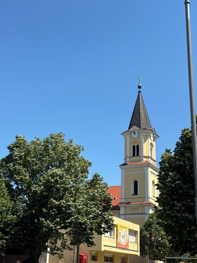 The image captures the steeple of a yellow church in Siófok, Hungary. The steeple has a clock face and a pointed roof. A metal pole runs along the top left of the frame. Part of a building is visible next to the steeple, adorned with a sign in Hungarian. Trees frame the steeple on the right. The sky is clear and blue.