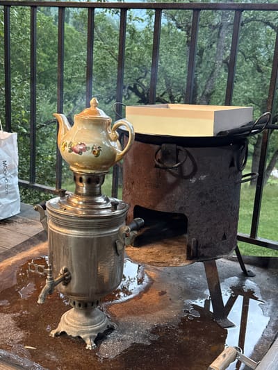 An outdoor scene on a balcony or porch shows a traditional samovar next to a rustic metal stove, with a wooden box on top of the stove. The samovar is made of polished metal and has a decorative ceramic teapot on top, adorned with painted fruit. A spigot is visible on the side of the samovar. The metal stove is dark and appears weathered, with an opening for fire at the bottom. The wooden box is light-colored and rectangular. The ground beneath the objects is wet and appears to be concrete or a similar surface, with some puddles reflecting the surroundings. In the background, a railing separates the balcony from a lush green, wooded area, suggesting a natural, possibly mountainous, environment. There are no people visible in the image. The overall impression is of a setup for preparing tea outdoors, possibly in a rural or scenic location. While the prompt asks for the location in Sis, Azerbaijan, there are no visual cues in the image that specifically identify it as such.