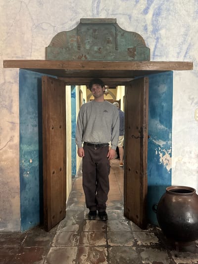 A man in a gray sweater and brown pants stands framed in a weathered wooden doorway. The doorway is flanked by a decorated, blue-painted wall on one side and a blue and white wall with a large brown pot on the other. The floor is tiled in gray and brown. Another person is faintly visible in the background through the doorway.