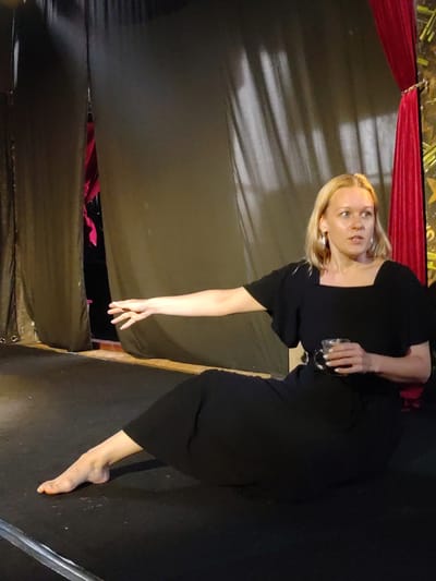 A Caucasian woman with blonde hair is seated on a black surface, barefoot and wearing a black dress. She extends her left arm, gesturing outward with an open hand. In her right hand, she holds a clear glass. Behind her are theatre curtains, including heavy black curtains and a red curtain on the right. The setting appears to be Bedulu, Indonesia, as the location was requested.