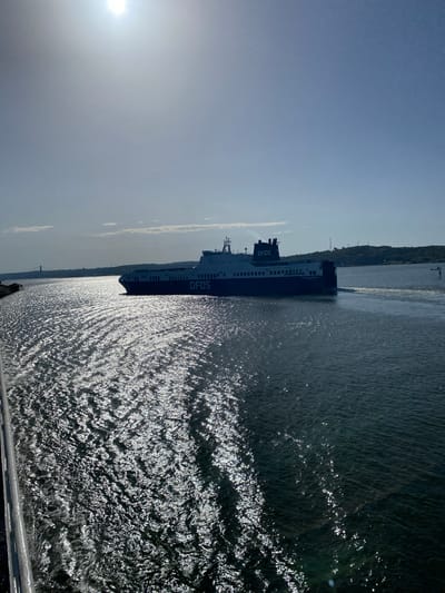 The media file depicts a DFDS ferry navigating through the water. The ferry is a large, dark vessel with multiple decks and windows. The name "DFDS" is clearly visible on the side of the ferry in white lettering. The water is choppy and reflects the sunlight, creating a shimmering effect. In the background, a tree-lined shore is visible, with some structures suggesting a coastal or port area. The sky is bright and clear, with a hazy sun in the upper left portion of the image. The overall impression is of a bright, sunny day. The image is oriented vertically, suggesting it was taken from a higher vantage point, possibly another vessel or a bridge. The location is identified as Gothenburg, Sweden.