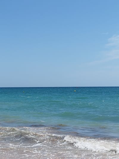 A wide, clear blue sky dominates the upper half of the image. The horizon is a sharp line dividing the sky from the ocean. The ocean is a vibrant turquoise color, with small waves and whitecaps indicating a moderate breeze. Two yellow buoys are visible in the water, with a small dark boat in the far distance. The foreground shows the edge of a sandy beach with small waves gently washing ashore, creating white foam. The overall scene depicts a sunny day at the beach in Rota, Spain.