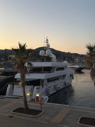 This media file captures a yacht docked in a harbor. The yacht is a large, white vessel with multiple levels, characterized by its sleek design and numerous windows. In the foreground, a palm tree with dried fronds stands in a planter. Behind the yacht, the water of the harbor is calm, reflecting the sky and the lights from a distant town nestled on a hillside. The town's architecture is visible, with many buildings clustered together, suggesting a Mediterranean coastal settlement.
The image appears to have been taken during the golden hour, with a soft, warm light illuminating the sky, transitioning from a pale blue at the top to a soft orange near the horizon. This suggests either sunrise or sunset. The weather seems clear and calm, with no indication of wind or rain.
The scene is set in Imperia, Italy, as indicated by the location context. The overall mood is serene and peaceful, evoking a sense of quiet luxury and the beauty of a coastal evening. There is some visible text on the side of the yacht, appearing to be the name "TORRILLE" with a stylized "T" above it. Additionally, there are life preservers and other dockside amenities visible near the yacht. The image is oriented vertically.