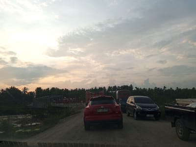 This is a photograph of a dirt road in Awegeutahpaya, Indonesia, at sunset. The sky is cloudy with the sun setting on the left, casting a warm glow. A red Nissan Juke is in the foreground, facing away from the camera. To its right is a black Toyota Avanza, and further to the right is a black pickup truck. In the background, a bridge spans a body of water, with a line of trees and palm trees beyond it. Several trucks are visible on the bridge, and flags are attached to some of them. The overall scene suggests traffic congestion on a rural road during twilight.