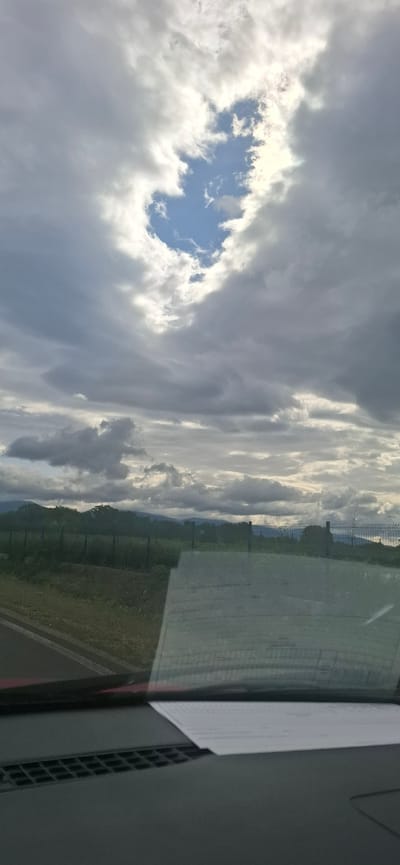 This image shows a view from inside a vehicle, looking out through the windshield. The foreground of the image shows the dashboard of the car, with a piece of paper resting on it. The windshield is visible, reflecting the sky and the landscape outside.
The sky is predominantly filled with dark, heavy clouds, with a bright patch of blue sky visible in the upper center, peeking through a gap in the clouds. This opening in the clouds is illuminated by sunlight, creating a contrast with the surrounding darker clouds.
Beyond the windshield, a fence is visible, made of metal posts and wire mesh. Behind the fence, there is a green field with some vegetation and trees. In the distance, rolling hills or mountains are visible, partially obscured by the clouds and haze. The overall atmosphere suggests an overcast or partly cloudy day, possibly before or after rain. The scene is devoid of people or any specific action. The location in the city of Dolno Sahrane, Bulgaria, cannot be determined from the visual information present in the image.