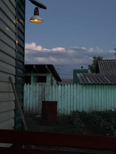The image depicts a twilight scene in Rodino, Russia. The sky is a deep blue with scattered clouds, suggesting the transition from day to night. The foreground features the roof of a building with a corrugated metal surface, reflecting the dim light. To the left, a security light with a bright yellow bulb is attached to a pole, illuminating the surrounding area.
Behind the foreground building, another structure is visible. It is a wooden building with weathered, pale green siding. The wood grain is clearly discernible, and some planks show signs of age. The roof of this building is covered with corrugated material, possibly asbestos-cement sheeting, which appears gray and weathered. Above this structure, another section of roofing with a similar corrugated material is visible.
To the right of the wooden building, dense green foliage grows, indicating a natural or overgrown area. A dark, rusty metal object, possibly a barrel or tank, is leaning against the wooden wall. Further to the right, a dark red vertical post, likely part of a fence or gate, occupies the edge of the frame, with a portion of a similar post visible on the far right. A wooden pole or handle is visible lying diagonally across the lower section of the image, resting on the corrugated roof.
The overall impression is one of a rural or semi-rural setting, possibly a backyard or an outbuilding area, during the evening. The lighting suggests the time is dusk or early evening, as the artificial light from the security lamp is on, and the sky is still somewhat bright. There is no visible text in the image. The scene evokes a sense of quietness and perhaps a touch of rustic melancholy.