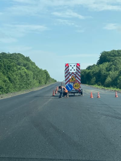 The image depicts a road construction site on a clear day. The road surface is dark asphalt, with some lighter patches indicating recent work. On the right side of the road, several orange traffic cones are placed at intervals. In the background, a large, portable traffic control device with flashing red lights and various road signs, including a construction worker symbol and a "40" speed limit sign, is positioned. A person wearing an orange vest and dark pants is seen working near this device. A dog is also present near the worker. The road is bordered by dense green trees on both sides, suggesting a rural or semi-rural setting. The sky is bright blue with scattered white clouds. The overall scene indicates ongoing road maintenance or construction work in what appears to be the vicinity of Zanadvorovka, Russia.