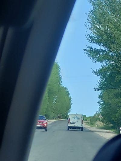 This image captures a daytime scene on a paved road, likely viewed from inside a moving vehicle. In the immediate foreground, a dark, blurry element, possibly part of a car's interior, partially obstructs the view.
On the road ahead, a white commercial van is prominently visible. The word "TATNI" is printed in blue on its rear door, beneath a blue logo resembling a triangular roof with a horizontal line. The van's rear lights are visible, and its license plate is partially obscured. Further down the road, behind the white van, another vehicle can be seen, appearing to be a red and blue truck or car, with its red taillights illuminated.
The road is flanked by lush green trees and foliage, extending into the distance under a clear, bright blue sky. Faint lines, possibly utility cables, are discernible crossing the sky between the tree canopies. The overall impression is one of clear weather during daylight hours. The scene is set in Zangakatun, Armenia.