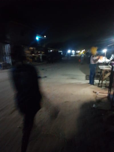 The image depicts an outdoor scene in Awoyaya, Nigeria, at night. The lighting is poor, with some lights visible in the background. The foreground shows a blurry, dark figure. In the mid-ground, a person wearing a yellow head covering is standing next to a table. There are other people and structures visible further down the road. The road appears to be unpaved.
