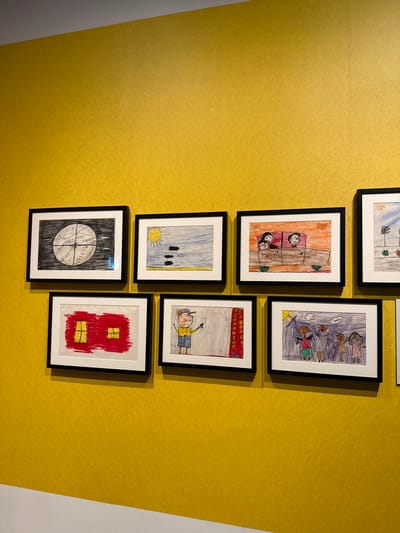 Seven framed children's drawings hang on a mustard-yellow wall. The frames are black with white mattes. From top to bottom and left to right, they depict winter, an interior scene, students walking, a sunny field with trees, a child on the floor, a dark circle, and a red building. The wall transitions to white in the lower right of the frame.