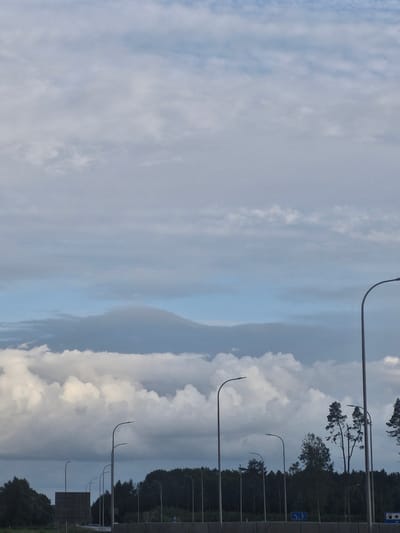 The image depicts a view from a road, likely a highway, with a line of streetlights and a dense forest in the background. The sky is filled with a dramatic cloudscape, featuring both wispy cirrus clouds at higher altitudes and large, puffy cumulus clouds closer to the horizon. A dark, layered cloud formation is visible in the mid-ground, adding depth to the sky. The trees in the background are dark green and form an unbroken line, suggesting a natural or park area. Several streetlights, with their distinctive curved arms, are spaced along the road. A large, dark, rectangular structure, possibly a billboard or sound barrier, is visible on the left side of the frame. The overall scene is overcast, with muted colors, and the lighting suggests it is either early morning or late afternoon. The location is identified as being in the vicinity of Jenči, Latvia.