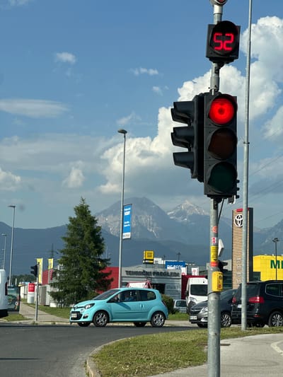The image captures a street scene in Kranj, Slovenia, dominated by a traffic signal displaying a red light and a countdown of 52 seconds. To the right of the traffic light, along the street, a light blue Renault Twingo is parked, with a license plate of KR-SF-969. Beyond the car, several commercial establishments are visible. Signs indicate a Toyota dealership and service center, as well as the presence of "BIG BANG", a "TEHNIČNI PREGLEDI" facility, a "FITINN", and a "STOP SHOP". Further down the street, there are more cars and some street lights. In the background, a mountainous landscape merges with a partly cloudy sky. The setting sun casts long shadows across the street. A sign visible in the background reads, "Produini center HOBBY", and "Ulica Mirka Vadnova 19a", helping to pinpoint the exact location of the scene.