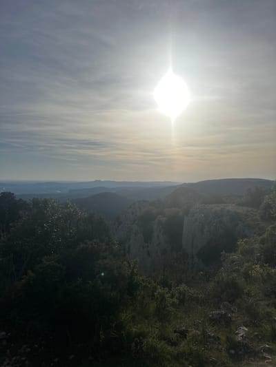 The image shows a landscape near Bonnieux, France. The sun is positioned high in the sky, dominating the left side of the frame with its bright light. Clouds are scattered thinly across the sky. To the right, a rocky cliff face descends, covered in vegetation. In the distance, there are layers of mountains fading into the hazy atmosphere. In the bottom of the image, vegetation obscures the view, which indicates that the image was taken from a high vantage point. There are no people present.