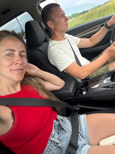 A man and a woman are in the front seats of a car, driving. The woman is in the passenger seat, facing the camera with a slight smile. She has long blonde hair and is wearing a red tank top and denim shorts. The man is in the driver's seat, looking forward and driving. He is wearing a white t-shirt and olive green shorts. The car's interior is visible, including the dashboard, center console, and car seats. The scene appears to be a daytime drive on a highway, with trees and fields visible through the car windows. The location is specified as Novoe Mart'yanovo, Russia.