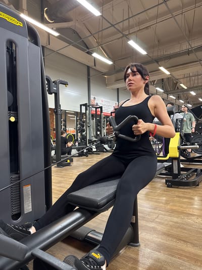 In an indoor gym, a woman with dark hair tied back in a ponytail is seated on a weight machine, performing a rowing exercise. She is wearing a black sleeveless top and black leggings. Her hands grip a black cable attachment, and a red bracelet is visible on her left wrist. The gym has a wooden floor, and various weight machines and equipment are visible in the background, including a yellow machine and racks of weights. Another person, a man wearing a green t-shirt and black shorts, is standing in the background. The gym appears to be equipped with fluorescent lighting and industrial ventilation systems visible on the ceiling. The gym is located in the city of Chaikovsky, Russia.