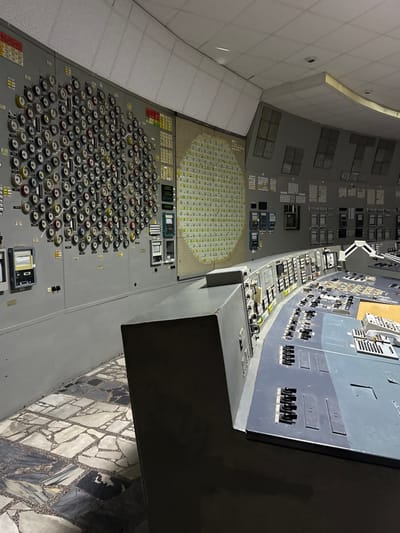 This image depicts the Control Room of Reactor 4 at the Chernobyl Nuclear Power Plant, located near Pripyat, in the Kyiv Oblast, Ukraine. The scene is devoid of human presence, presenting an aged industrial control environment.

The left wall features extensive grey control panels. The leftmost panel segment is covered by a dense array of circular gauges and switches, arranged in an irregular grid-like pattern. Adjacent to this, a large, circular, illuminated mimic display dominates the central portion of the wall, presenting a grid of smaller, uniformly sized rectangular lights or indicators, predominantly in shades of green and yellow, likely representing a reactor core diagram. Further to the right, additional vertical panels with various meters, screens, and rectangular buttons are visible.

In the foreground and extending into the midground, a long, curved control console, primarily dark grey, occupies the space. Its surface is replete with an assortment of black switches, buttons, and smaller rectangular displays. The console's design suggests multiple operator workstations.

The floor is covered with large, irregularly shaped tiles in a mosaic pattern of light grey, dark grey, and black, exhibiting a worn appearance. The ceiling features light-colored, possibly acoustic, panels. The lighting is functional but appears somewhat dim, casting subtle shadows within the room. No people, objects in motion, or interactions are present.