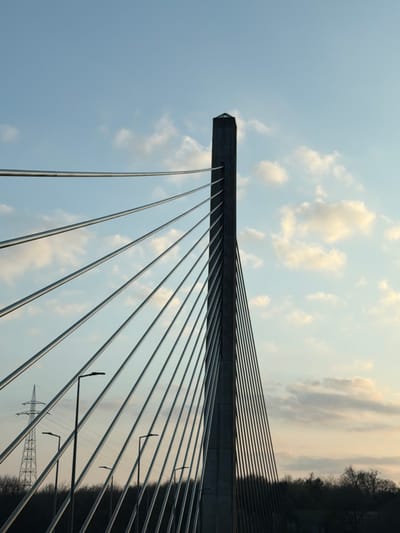 The image displays a close-up, low-angle view of the central pylon and tension cables of a cable-stayed bridge, identified as the Pont Victor Bodson located near Itzig, Luxembourg. The prominent feature is a tall, slender, dark gray concrete pylon situated on the right side of the frame, extending vertically towards the top. Numerous parallel, silvery-gray cables radiate diagonally downwards and to the left from the pylon, creating a fanning pattern across the upper and middle sections of the image. The sky, occupying the upper portion, is light blue with scattered white and faint orange-pink cumulus clouds, indicative of late afternoon or early evening light. Below the cables, the silhouettes of at least five modern streetlights with flat, horizontal fixtures are visible. In the distant background on the far left, a tall, lattice-structured electricity pylon is discernible against the sky. The lower edge of the image is defined by a dark, silhouetted tree line or dense vegetation. No individuals or vehicles are present in the frame.