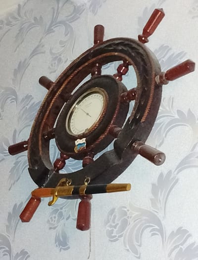 The image displays a decorative wall clock shaped like a ship's wheel. The wheel is made of dark wood with a rope trim around its outer edge. It has several wooden handles protruding from its spokes. In the center of the wheel is a clock face with Roman numerals and a single hand, indicating the time. Below the clock face, there is a small emblem that appears to be a flag with blue and white stripes and a red stripe, possibly representing the Ukrainian flag. Attached to the bottom of the ship's wheel is a decorative object resembling a miniature sword or dagger with a black handle and a golden hilt, further enhancing the nautical theme. The clock is mounted on a light blue wallpaper with a subtle floral or damask pattern. The lighting suggests it is daytime, but no specific time of day can be determined from the image. The setting appears to be indoors, likely a home in Hamarnia, Ukraine, given the provided location context. There is no discernible activity or event taking place, as the image focuses solely on the decorative clock.