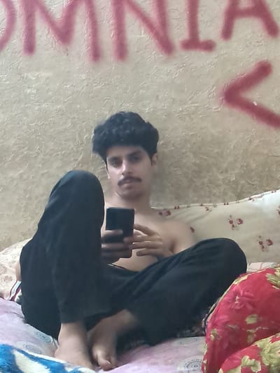 The image shows a young person lying down, holding a smartphone. The person has dark, curly hair and appears to be looking at the phone. They are wearing black pants and a black top. The background consists of a textured wall with some graffiti or spray paint in pink and red. A patterned blanket or bedding is visible behind the person. The scene appears to be indoors, possibly in a bedroom. There are no clear indicators of the time of day or weather conditions. The overall mood is casual and relaxed. The spray paint on the wall contains some visible text, though it is largely obscured and difficult to decipher completely. However, fragments of letters like "V" and "TA" are partially visible. The location context provided is Abbottabad, Pakistan.