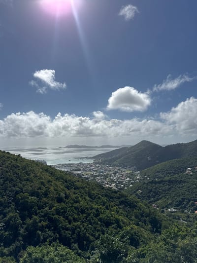 A landscape view features a town nestled between lush green mountains and a body of water. The town appears dense with buildings, sloping towards the water's edge. The sky above is a bright blue with scattered clouds. The sunlight streams in from the left, creating a lens flare effect. The vegetation on the mountains is thick, covering the slopes.