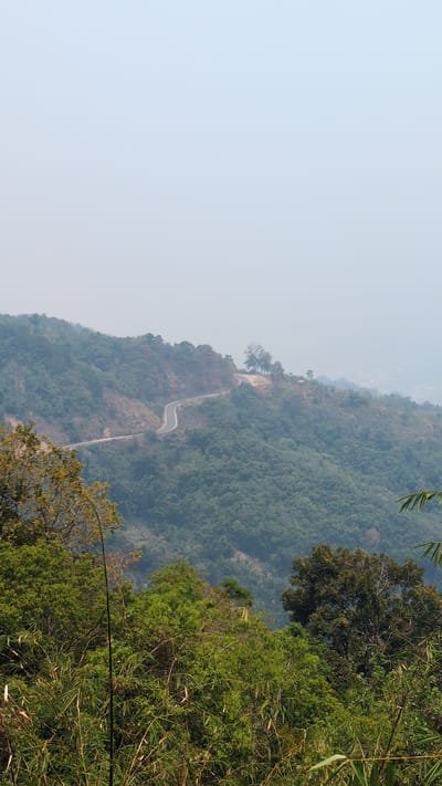 The image depicts a mountain landscape in Lâm Sơn, Vietnam. A winding road ascends the side of the mountain. The terrain is covered in dense green vegetation. The sky is hazy and light blue.