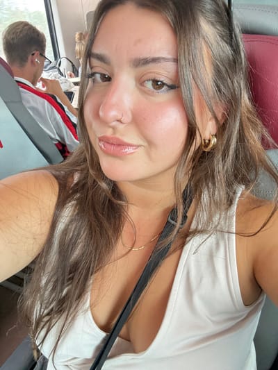 This image depicts a selfie taken by a female individual inside a public transportation vehicle, likely a train, in the vicinity of Castelfranco Emilia, Italy. The primary subject, a young adult female, occupies the foreground, facing the camera with a subtle smile. She presents with light skin, long brown hair, and dark eyes, enhanced by dark eyeliner and shiny, light pink lip gloss. She wears a small silver nose ring on her left nostril and gold hoop earrings. A delicate gold chain necklace is visible around her neck, above a light-colored tank top. A black strap, possibly from a bag, crosses diagonally over her chest. In the mid-ground, to the left, a male individual is partially visible from the back-left shoulder. He has short light-colored hair, wears white wireless earbuds, and a white shirt with red and black striped sleeves, appearing to look downwards. The environment consists of grey passenger seating with red headrests, and a window on the left reveals blurred green vegetation, indicative of a moving conveyance.