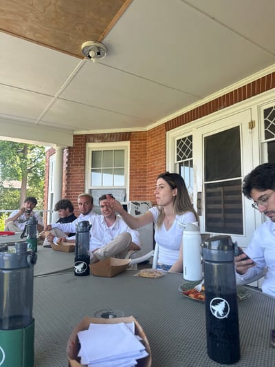 This media file depicts a group of people gathered on a porch, likely during a daytime social event. The scene is outdoors, with a brick building and a white-columned porch visible in the background, suggesting a residential setting. A table with a textured surface occupies the foreground, covered with various items including food, drinks in water bottles, and paper.
The subjects are several individuals, some of whom are wearing white shirts. They appear to be interacting with each other and the objects on the table. One woman in a white t-shirt is looking down, holding a water bottle. A man with glasses and a white shirt is looking towards the camera. Another man is gesturing with his hand while looking at the camera. The overall mood seems relaxed and casual.
Notable details include several reusable water bottles, one black with a white logo of a tree and a bird, and another white with text that reads "owala." There are also brown paper bags, some containing food items and others filled with white paper. The time of day appears to be daylight, and the weather seems pleasant, conducive to an outdoor gathering. The location cues, such as the architecture of the building and the porch, suggest a possibly suburban or historic setting in the United States. The individuals' expressions and interactions suggest a friendly and informal occasion.