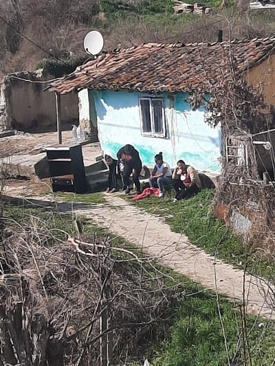 The image depicts an outdoor scene in Ivanski, Bulgaria, featuring a light blue painted house with a reddish-brown tiled roof. A white satellite dish is mounted on the roof. In front of the house, a concrete platform holds a dark, rectangular object and a plastic bottle. Four individuals are gathered on the ground level in front of the house.
Two individuals, both dressed in dark clothing, are bent over near the center of the frame. A red fabric item lies on the ground directly in front of them. To their right, two other individuals are seated. One individual, wearing a white top and light-colored pants, is looking towards the bent individuals. The fourth individual, wearing a reddish-brown top, has their head resting on their left hand and is looking downwards.
The immediate foreground consists of dense, bare branches of shrubs or trees, indicative of dormant vegetation. A light-colored path curves through green grass from the left mid-ground towards the right foreground. The background reveals a sloping hillside with sparse, dry vegetation.
