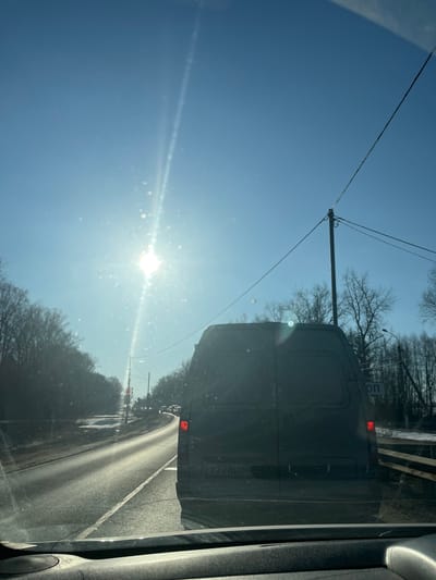 The image depicts a daytime view from the interior of a vehicle traveling on a multi-lane asphalt road in Rogachovo, Russia. A clear, bright blue sky with a prominently visible sun in the upper-left quadrant generates significant lens flare and glare on the observer's windshield, which also shows multiple bright spots consistent with dust or debris. Directly ahead, a dark-colored commercial van, possibly grey or black, occupies the same lane. Its rear is visible, including an illuminated red taillight on the right side and a partially obscured Russian license plate beginning with "T778BO." The road surface appears reflective under the sunlight, with visible white lane markings. Bare deciduous trees line both sides of the road, and patches of snow are visible on the shoulders and adjacent ground. Utility poles with overhead lines are present along the right side of the road, where a white sign with Cyrillic text, including "ОП," is partially visible behind the van. Other vehicles are faintly discernible in the distance down the road. The top of the observer's vehicle dashboard is visible in the lower foreground. No people are explicitly visible.