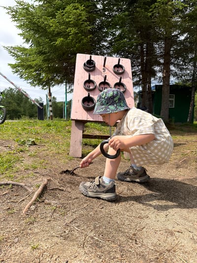 A young boy wearing a bucket hat and a checkered shirt and shorts crouches in a dirt and gravel area. He holds a ring in his right hand and appears to be digging with a small trowel in the dirt. Behind him, a pink wooden board with nine black rings and pegs stands upright. Trees and a green building are visible in the background. A white and red barrier gate is also present to the left of the frame. The setting appears to be an outdoor recreational area, possibly in Kul'dur, Russia, given the mention of the city, though no specific identifiers are present within the image itself to confirm this location.