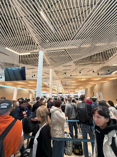 The image depicts a crowded airport terminal in the city of Ianeti, Georgia. Many people are standing in lines, likely waiting to check in or go through security. Some have luggage. The terminal features a modern design with a prominent white, patterned ceiling and a large screen displaying information. The overall impression is one of a busy and modern airport.
