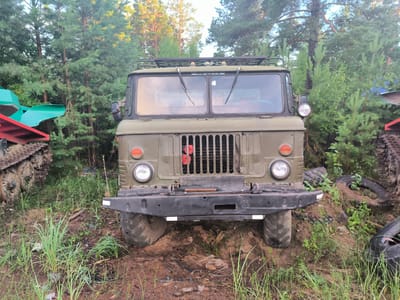 A green military truck, likely a GAZ-66, is parked in a wooded area, possibly the outskirts of the village of Gremiuchiy, Russia. The truck's windshield is cracked, and the headlights and grille are visible. To the left of the truck, a portion of a green and red piece of machinery with tracks is present. The ground is covered in dirt, grass, and debris, suggesting an outdoor, possibly abandoned, setting. The background consists of dense trees, indicating a natural environment.