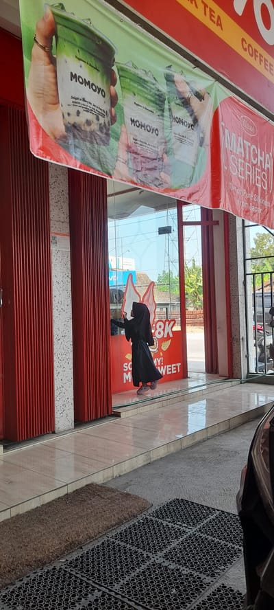 The image shows the exterior of a shop in Crewek, Indonesia, featuring a red retractable door and a glass storefront. A banner above the storefront advertises "MOMOYO" drinks and the "MATCHA SERIES." Inside the shop, visible through the glass, a young person wearing a black hijab and dress is interacting with a display featuring large cartoon ice cream cones and promotional text that includes "8K" and "SO YUMMY! MI SWEET". The ground outside the shop is tiled, with a textured mat and a coir mat on the left. A car is partially visible in the lower right corner. The lighting suggests it is daytime.