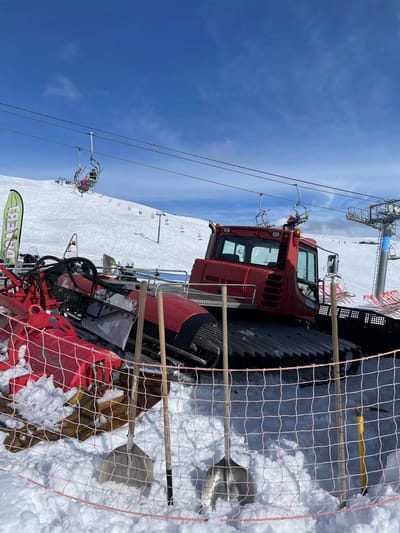 The image displays a sunny, snow-covered mountain landscape in Puy-Saint-André, France, characteristic of a ski resort. In the foreground, a red mesh safety net delineates an area where three shovels and one pickaxe are stuck upright in deep snow. Immediately behind this barrier, a large, red tracked snow groomer, identifiable by its cab and rear track system, is parked on the snow. To the left of the main groomer body, an additional red snow groomer attachment, likely a front blade or a grooming implement, rests on the snow.
Overhead, a multi-chair ski lift system spans the scene, with several chairs visible along its cables. At least two individuals are observed riding separate chairs; one person in an orange helmet is visible in a chair approaching the right, while another person in a pink jacket is seen in a chair further to the left, ascending the slope. Both individuals appear to be equipped for skiing or snowboarding, though specific equipment details are limited. The background reveals vast, groomed snow slopes dotted with more ski lift infrastructure and indistinct figures of other skiers. The sky is largely clear blue with minimal scattered clouds.
