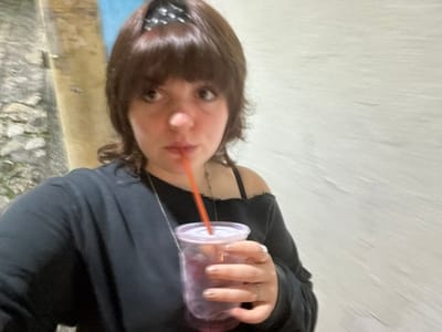 A woman drinks from a clear plastic cup with an orange straw. She wears a black top and a bandana in her hair. Her gaze is directed at the camera. The background is blurred, showing a textured wall. The image has been rotated so that the woman's head is at the bottom. The photograph appears to have been taken outdoors.