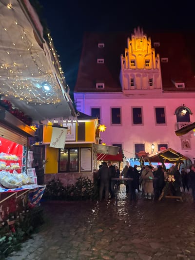 A nighttime outdoor shot shows a Christmas market scene. A building stands tall with brightly lit windows. Market stalls are decorated with lights and offer food like bratwurst. People are milling about. The ground is wet, reflecting light. The photo is taken from a slightly low angle.