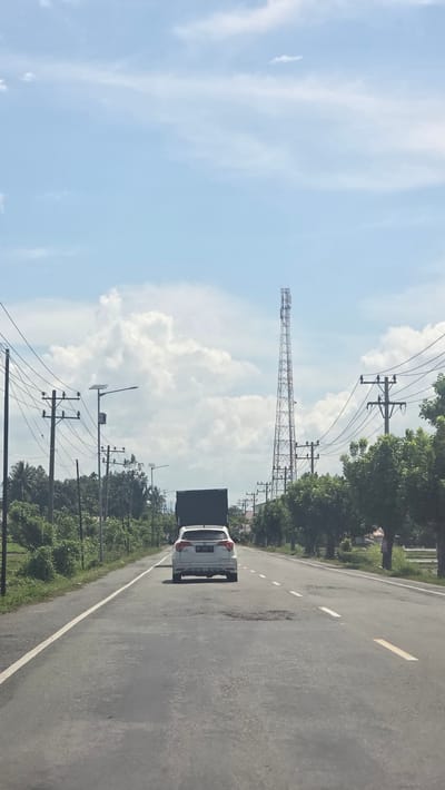 A street in Kampungjumpa, Indonesia. A white car travels on the right side of the road. Power lines run along the side of the road. In the background, a radio tower stands near cumulus clouds.