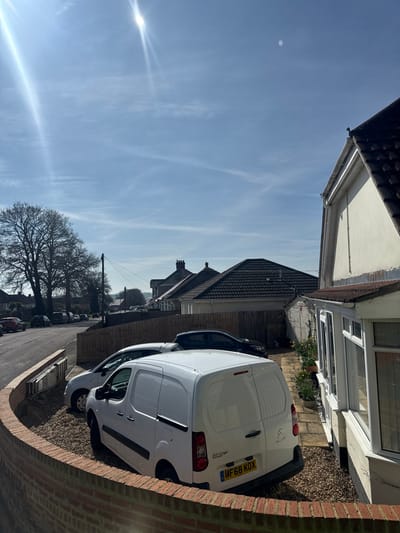This image captures a static residential scene in Salisbury, United Kingdom, on a bright, sunny day with a clear blue sky and sparse, wispy clouds. The perspective is slightly elevated, looking over a foreground brick wall into a gravelled driveway and towards a street lined with houses and trees.
In the immediate foreground, the top of a curved, reddish-brown brick wall is prominent. Beyond this wall, a gravelled driveway occupies the midground, featuring three parked vehicles. The most prominent vehicle is a white Citroen Berlingo panel van, licence plate MF68 KOX, parked with its rear facing the camera. To its left, a silver or light grey hatchback is partially visible. Further back, a dark-coloured car is also parked on the gravel.
To the right of the driveway, a cream-coloured house with a dark tiled roof and a white UPVC conservatory or extension is visible. A narrow paved path runs alongside this structure, where potted plants with yellow flowers (likely daffodils or narcissus) are arranged. A light brown wooden fence separates the driveway area from the properties beyond.
The background reveals a residential street curving to the left, lined with additional parked cars. Several large deciduous trees with bare branches stand prominently along the left side of the street. Beyond the street and neighbouring houses, a distant horizon with subtle landscape features is visible. Overhead utility poles and power lines traverse the scene above the street and buildings. No individuals are present, and no actions or interactions are depicted.