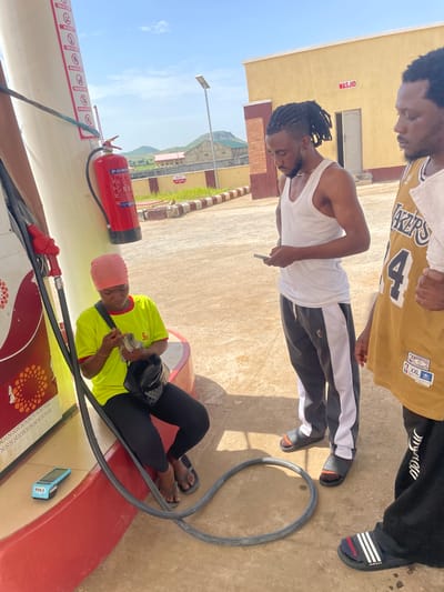 The image shows three people at a gas station in Pasere, Nigeria. One person, wearing a yellow t-shirt and a pink headwrap, is sitting and holding money, possibly preparing to pay or having just received change. They are next to a gas pump and a red fire extinguisher. Another person, wearing a white tank top and black and white striped pants, is standing and holding a small object, possibly a phone. They are also near the gas pump. The third person, only partially visible, is wearing a yellow and brown basketball jersey. The scene appears to be outdoors, with a sandy ground and a building in the background. The sky is clear and blue, suggesting it is daytime and likely sunny. Text visible in the image includes "POS 4", "TURN OFF BO", "NO", "POWDER", "BRYK", "MASJID", "LAKERS", "4", "HARDWOOD CLASSICS", and "XXL". The overall mood seems casual.