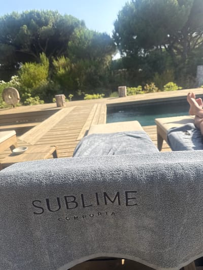 The image depicts a poolside scene at a resort in Grândola, Portugal. In the foreground, a gray terrycloth towel with the embossed words "SUBLIME" and "COMPORTA" rests on a lounge chair. Beyond the towel, a wooden deck surrounds a swimming pool. Two lounge chairs are visible on the deck, one covered with a gray towel and the other with a person's legs and feet visible, suggesting they are relaxing. Trees and foliage line the background, indicating a natural, outdoor setting. The lighting suggests it is a sunny day.