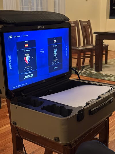 A POGA gaming case displays a soccer video game menu. "HOME" shows CA Osasuna from Spain's LaLiga with player stats, while "AWAY" features Liverpool from England with corresponding stats. The game appears to be in "UCL Final" mode. The portable monitor is angled and the case is open, revealing the interior foam padding. Part of a wooden chair is visible in the background.