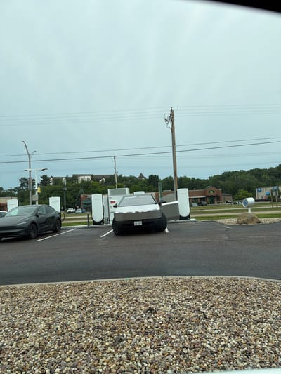 The image depicts a Tesla charging station in Wisconsin Dells, United States of America. In the foreground, a dark-colored Tesla Model Y is parked to the left, connected to a charging station. To the right, a silver Tesla Cybertruck is parked in front of another charging station, with its charging cable not visibly connected. Both vehicles are facing forward.

Behind the charging stations, several businesses are visible, including a Starbucks and a Jimmy John's. The environment appears to be overcast, with a gray sky. A variety of trees and buildings are present in the background, suggesting an urban or suburban setting. The foreground is dominated by a gravel area. The image appears to have been taken from a vehicle, as evidenced by the reflection on the lower part of the frame. No people are visible in the image.