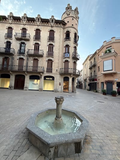 The image depicts a public square in Igualada, Spain, featuring a central octagonal stone fountain with a simple pedestal and basin, filled with water. The square is paved with light-colored rectangular bricks arranged in a diagonal pattern. In the background, a multi-story building with ornate architectural details stands prominently on the left. This building exhibits a curved corner tower with decorative circular motifs and crests, as well as multiple balconies with wrought-iron railings and dark wooden shutters on its windows. The ground floor of this building houses commercial storefronts, including a "Massimo Dutti" store with illuminated yellow signage. To the right of this building, a narrower street extends, flanked by other buildings of varying heights and colors, one of which is a salmon-pink building with a prominent bay window. The sky above is a clear, light blue with a few wispy clouds. No people are visible in the scene. The overall impression is of a quiet urban setting during the daytime.