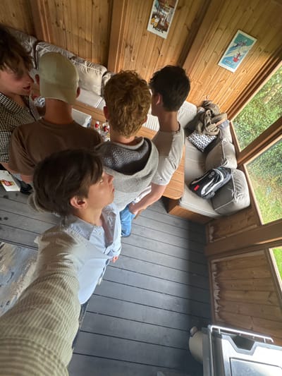 The image depicts the interior of a wooden outdoor structure located in Bubwith, United Kingdom. Five individuals, appearing to be males in their youth or young adulthood, are present. Four individuals are positioned with their backs or sides oriented towards the camera, standing near an L-shaped bench furnished with light grey cushions and a low wooden table. The fifth individual, occupying the immediate foreground, is capturing the scene from a low angle.
On the table, several clear bottles with red caps are visible. The wooden wall paneling features two framed posters, both displaying the word "MEGEVE" alongside ski resort imagery. A folded green blanket, a black and white patterned cushion, and a black sports bag with white and red accents are situated on the cushioned seating, the bag's shape consistent with a ski boot carrier. The floor of the structure is composed of dark grey slatted decking. Large glass windows line one side, offering a view of dense green foliage outside. A portion of a metal outdoor appliance is visible in the lower right foreground.