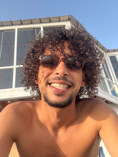 A close-up, chest-up selfie of a man with curly dark brown hair, sunglasses, and a beard. He is smiling, showing his teeth, and his shoulders are bare and glistening with water droplets, suggesting he is outdoors and possibly has been swimming. He is standing in front of a white building with large windows and a corrugated roof, possibly a beachside cafe or accommodation. The sky is clear blue and the lighting indicates it is daytime. The man's skin is tanned. The scene is consistent with the sunny coastal environment of Taghazout, a popular surfing destination in Morocco known for its beaches and relaxed atmosphere.